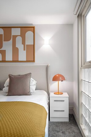 Een netjes ingerichte hotelkamer met een bed, een nachtkastje met een lamp, een kunstwerk aan de muur en een raam met zonwering.