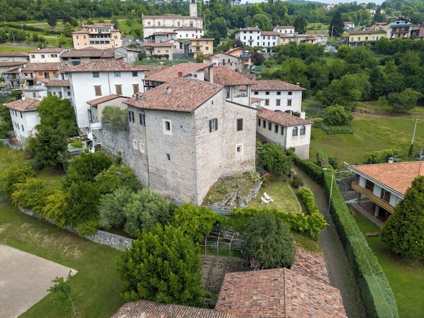 A stone castle with a tiled roof surrounded by lush greenery and residential buildings.