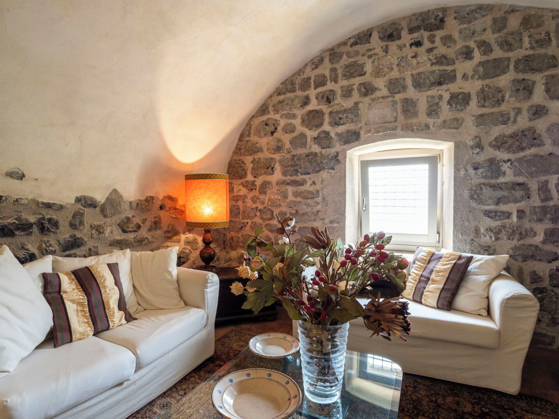 Une pièce chaleureuse avec un mur en pierre, deux canapés blancs ornés de coussins à motifs, une table sur laquelle est posé un vase de fleurs et une lampe.