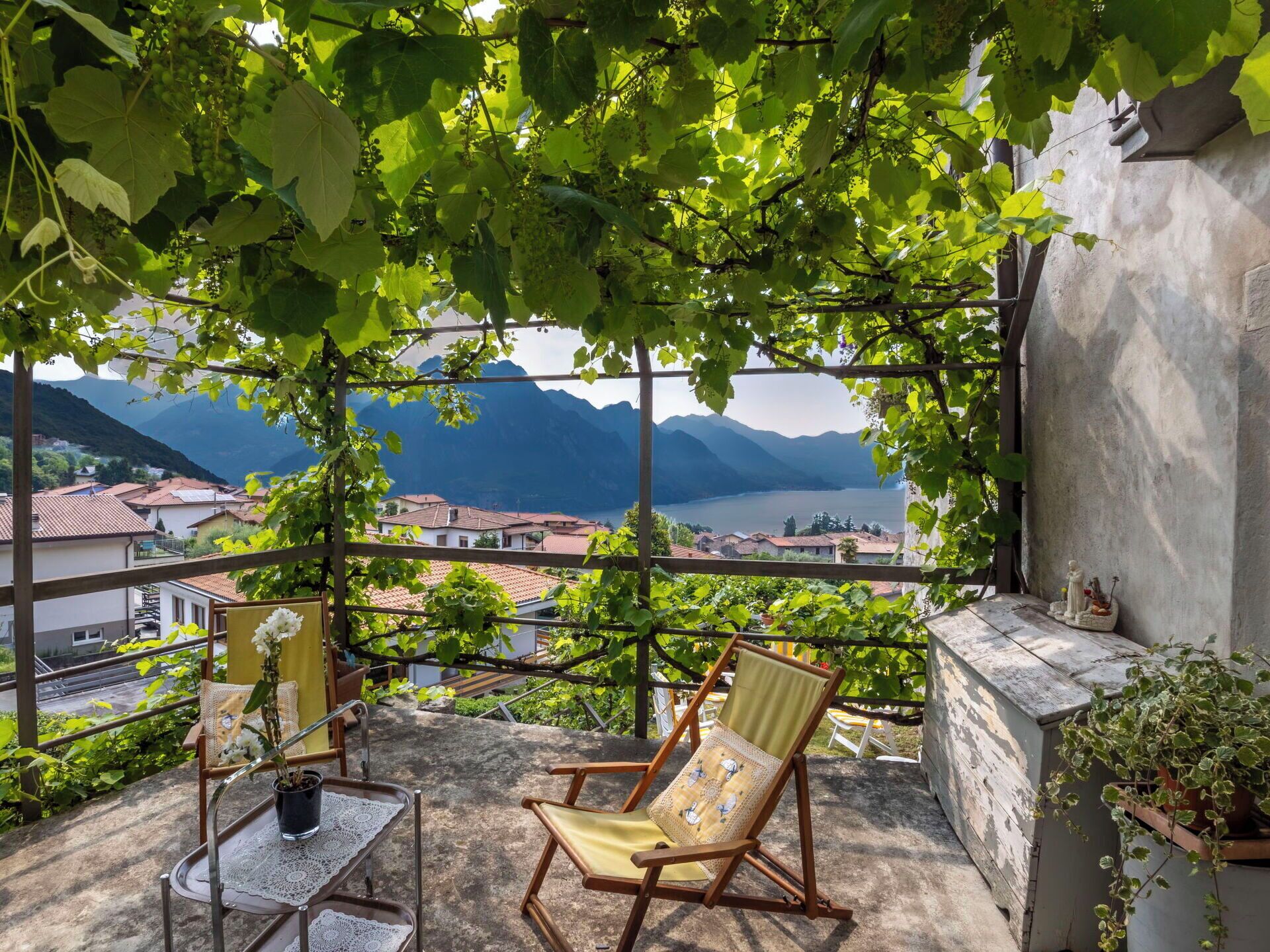 Ein Balkon mit Blick auf Berge und einen See, ein Holzstuhl, ein kleiner Tisch mit Spitze und ein Blumentopf.
