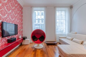 A modern living room with a red and white patterned wall, a flat-screen TV, a red chair, a white sofa, and large windows. - The Fitzroy - by Frankie (London)