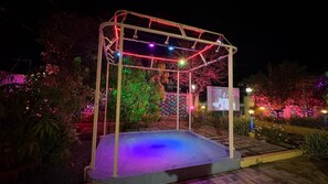 A nighttime outdoor area with a metal structure, possibly a gazebo, surrounded by illuminated plants and a small pool. - Cozy 1-bedroom farmhouse in serene Badlapur with AC (Badlapur)