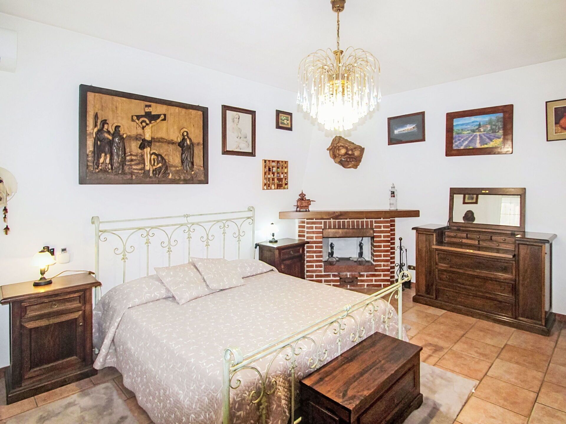 A bedroom with a bed, a fireplace, a dresser, a nightstand, and a chandelier.