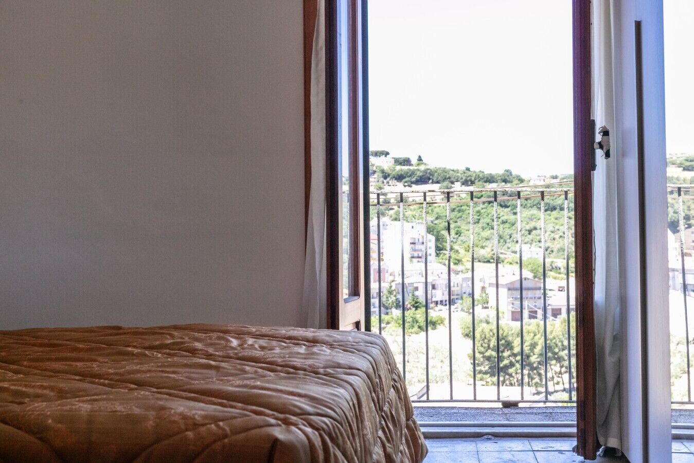 A bedroom with a bed, a balcony with a metal railing, and a view of buildings and greenery.