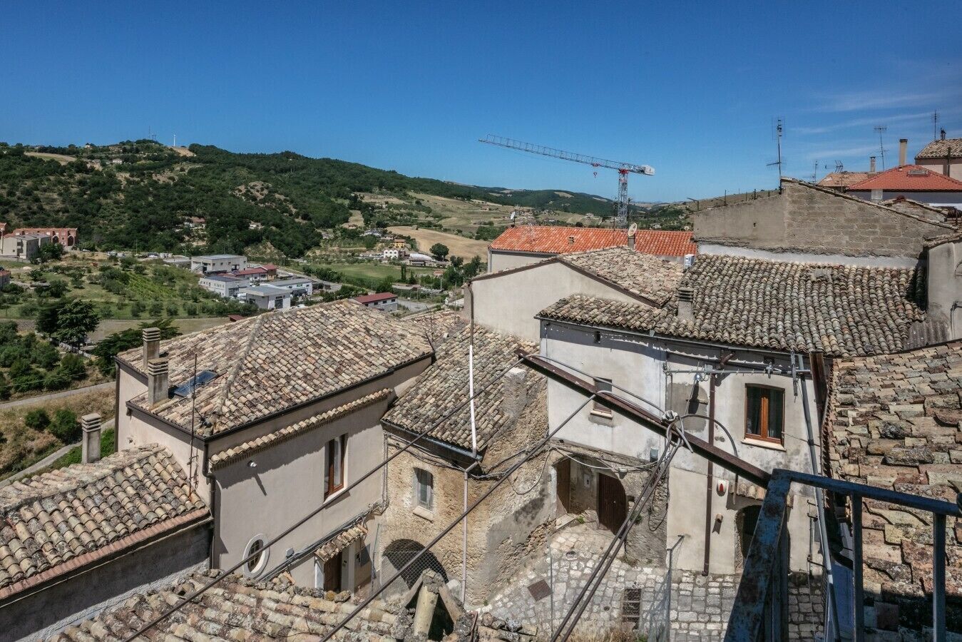 A view of a hillside town with terracotta-tiled buildings, a construction crane, and a clear blue sky.