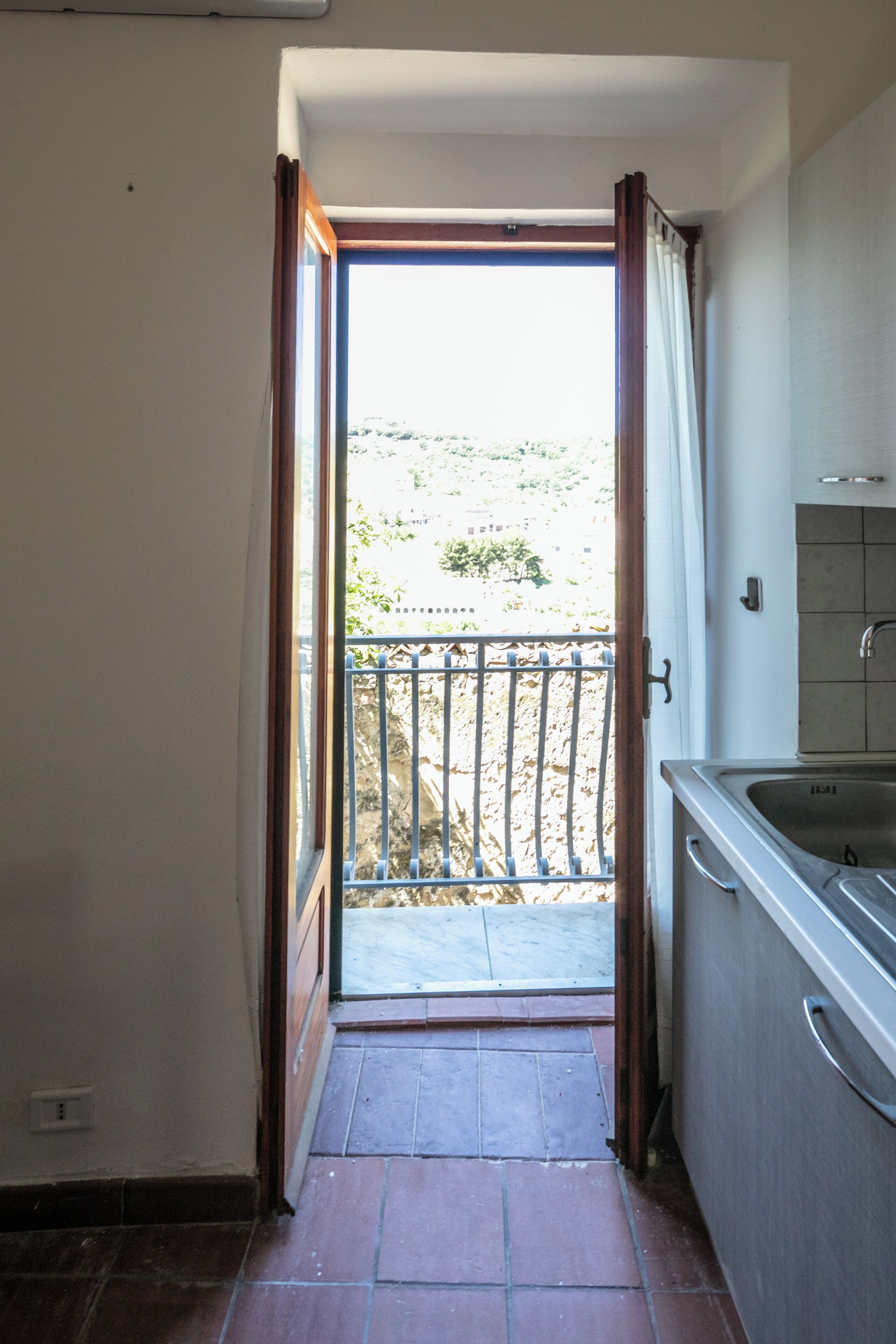 Een keuken met een spoelbak, een raam met gordijnen en een uitzicht op een balkon.