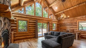 Interior de una cabaña de madera con chimenea, un sofá seccional y amplias ventanas que ofrecen vistas del exterior.