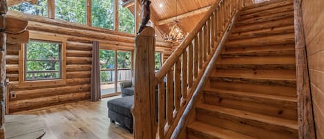 A log cabin interior with a staircase, large windows, and a chandelier.