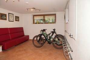A room with a red sofa, two bicycles, a window with lace curtains, and a wall-mounted bike rack. - Appartement Neumayer (Viehhofen)