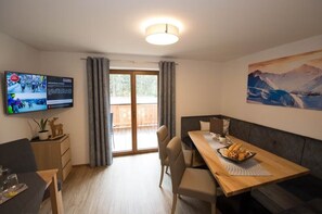 A modern room with a wooden dining table, chairs, a sofa, a TV mounted on the wall, and a painting of a mountain landscape. - Appartement Neumayer (Viehhofen)