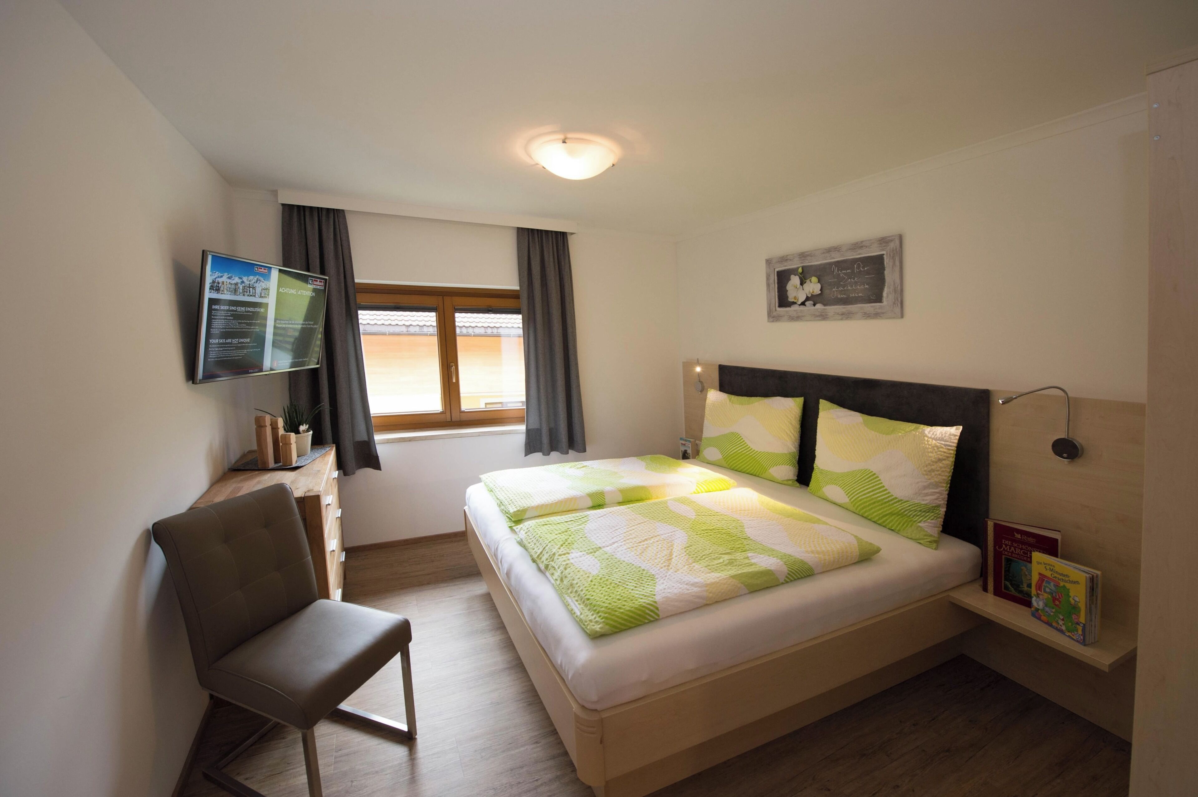 A bedroom with a bed, a chair, a desk, a TV, and a window.