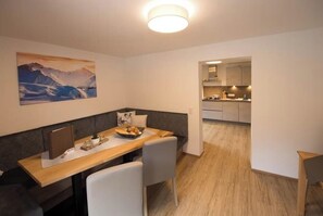 Un comedor con una mesa de madera, sillas blancas, un cuadro de montañas y una cocina al fondo.