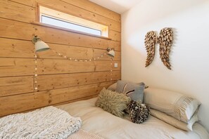 Quarto com paredes de painel de madeira, uma janela, duas luminárias fixadas na parede, uma cama com várias almofadas e um objeto decorativo na parede.