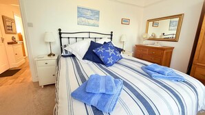 Ein ordentlich bezogenes Bett mit gestreifter Bettwäsche und Sternenkissen, ein Nachttisch mit Lampe, eine Kommode und ein Spiegel an der Wand.