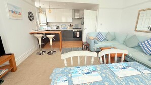 Ein modernes Wohnzimmer mit Sofa, Couchtisch und Barhocker. Im Hintergrund ist eine Küche mit weißen Schränken und einem Essbereich zu sehen.