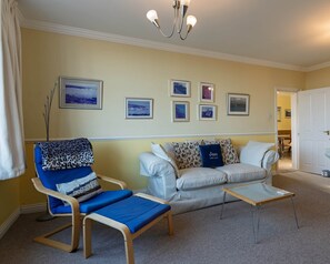 A living room with a blue armchair, a white sofa, a glass coffee table, and a chandelier. - Bright 2‑Bed Apartment with Balcony, Sea Views & Parking in Central Swanage (Swanage)