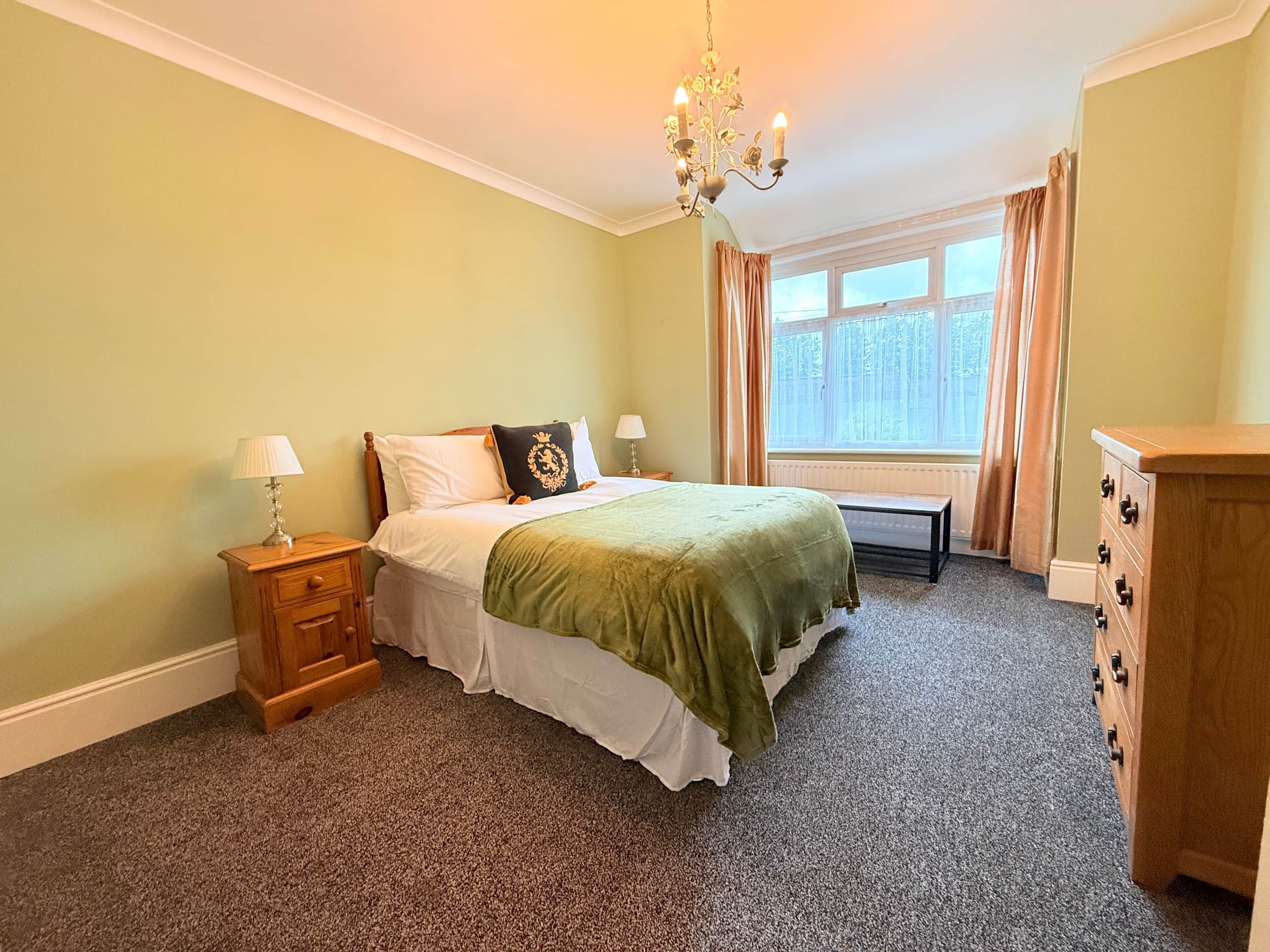 A bedroom with a bed, bedside tables, a dresser, a window with curtains, and a chandelier.