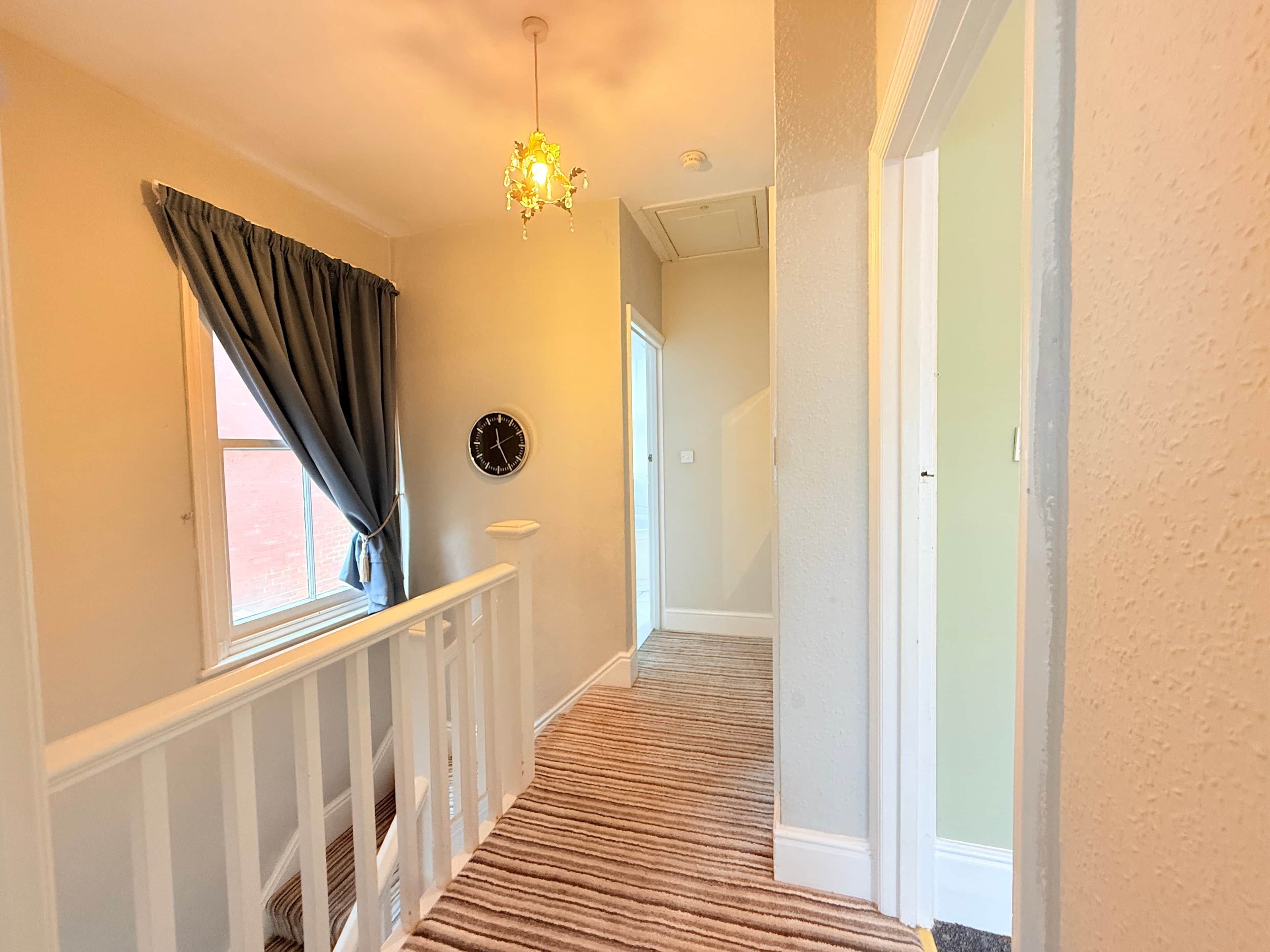 A hallway with a carpeted floor, a white railing, a window with curtains, a wall clock, and a door.