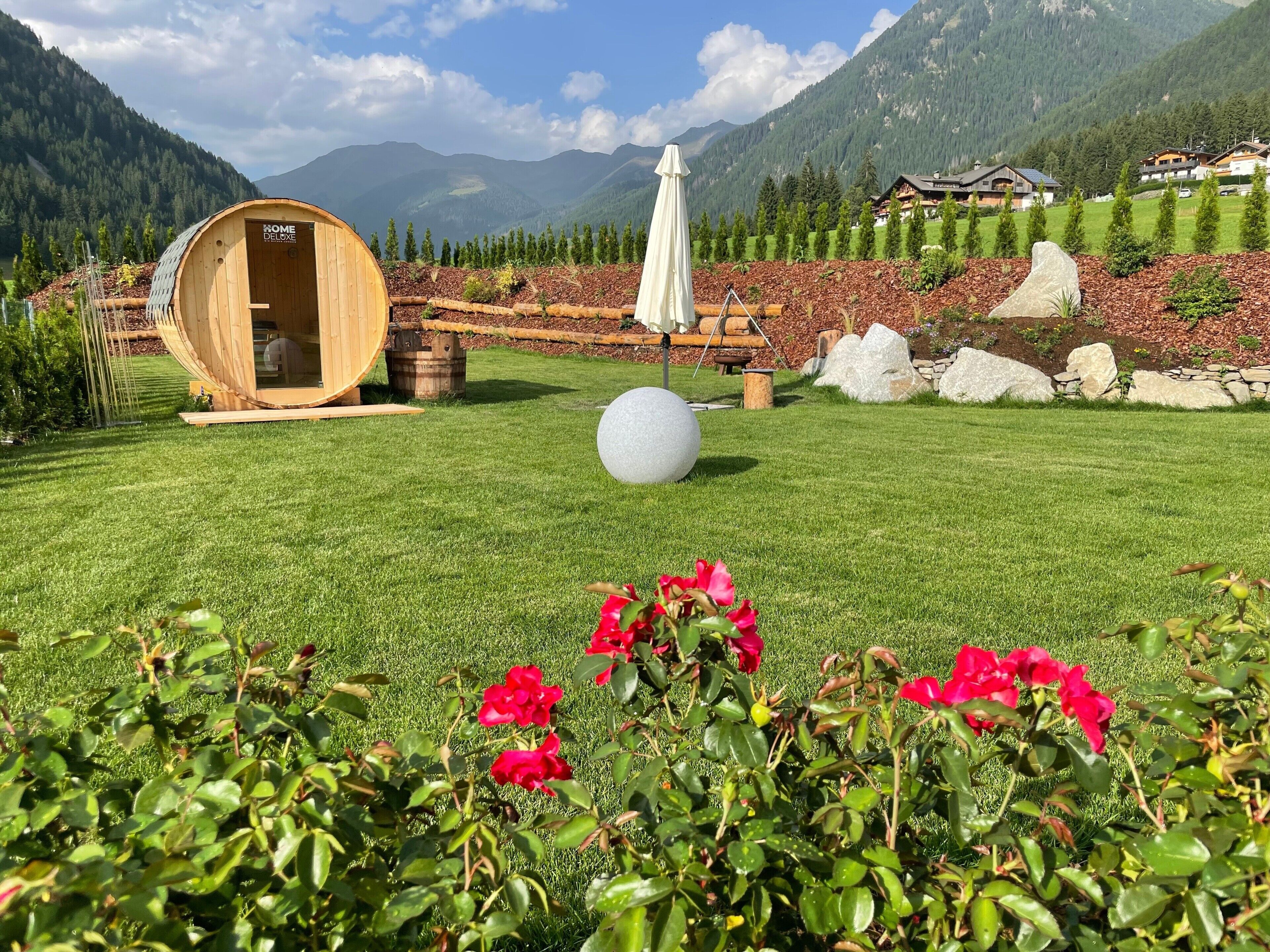 Una struttura sauna in legno, un ombrellone bianco e un oggetto sferico su un prato curato con fiori rossi in primo piano.