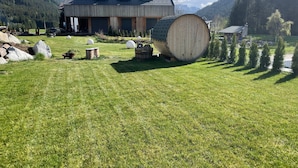 Una casa de techo inclinado, un barril de madera grande y un patio con césped, rocas y árboles.