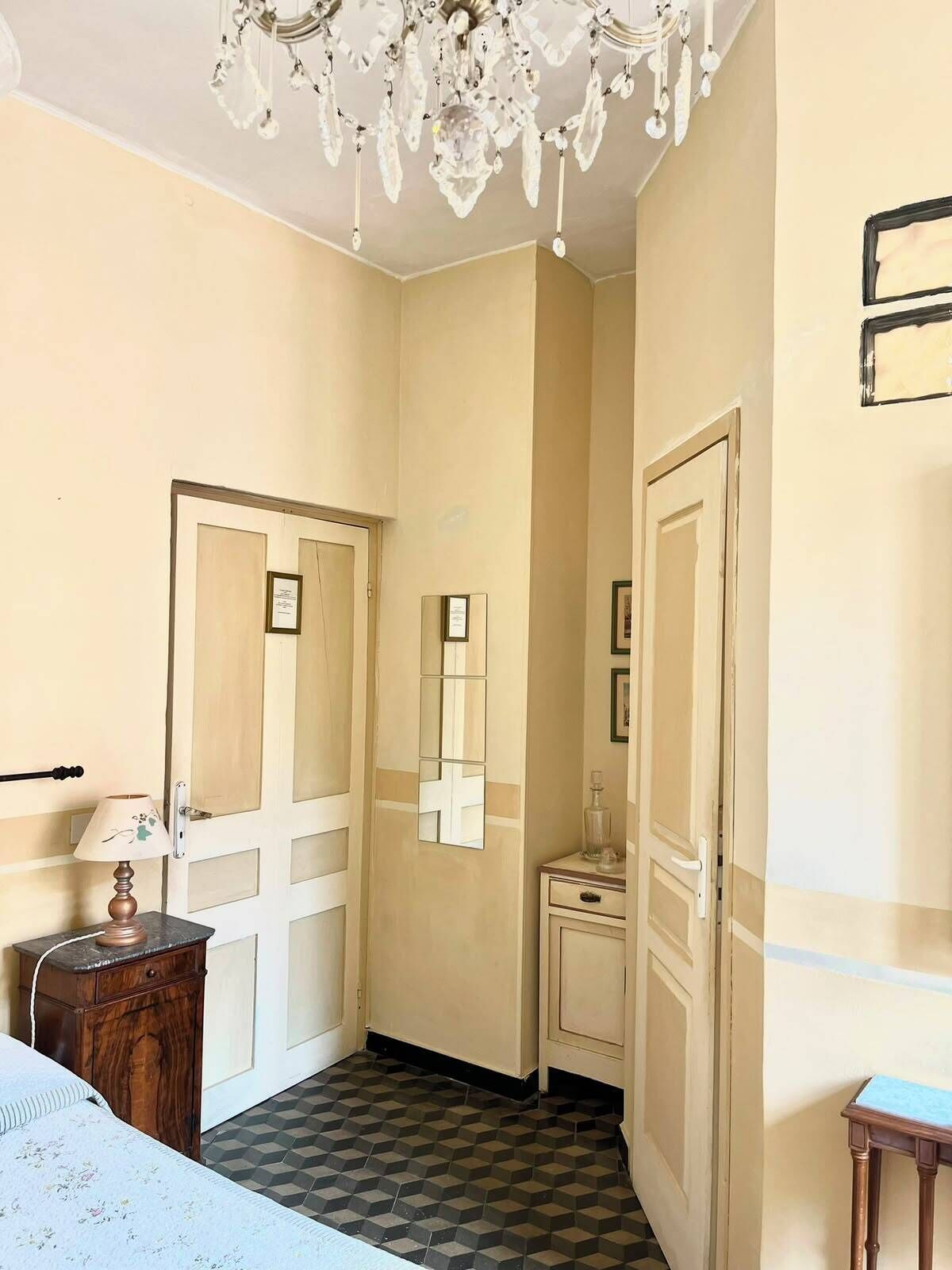 A narrow hallway with a chandelier, a bed with a floral bedspread, a small wooden nightstand, and a checkered floor.