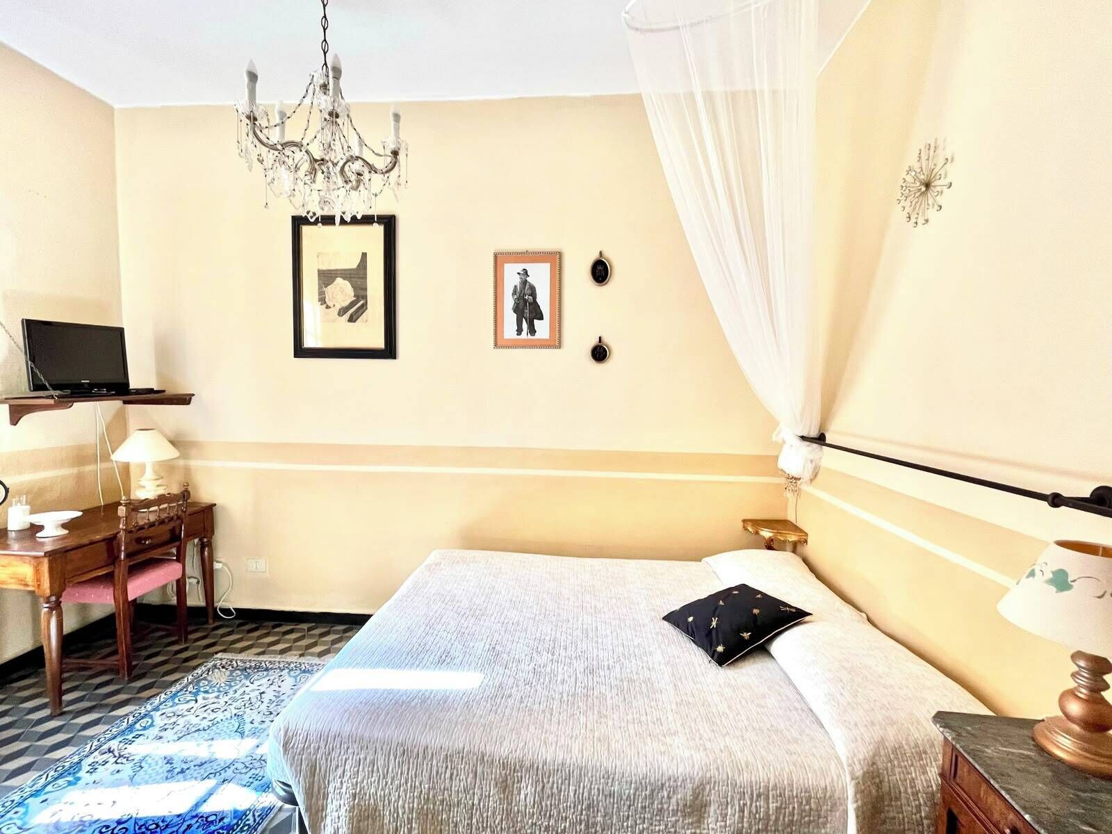 A hotel room with a bed, a desk with a lamp, a television on a shelf, and a chandelier.