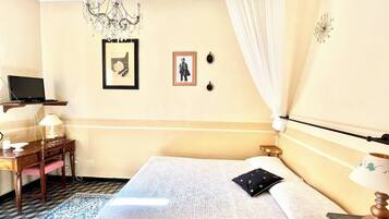 A hotel room with a bed, a desk with a lamp, a television on a shelf, and a chandelier.
