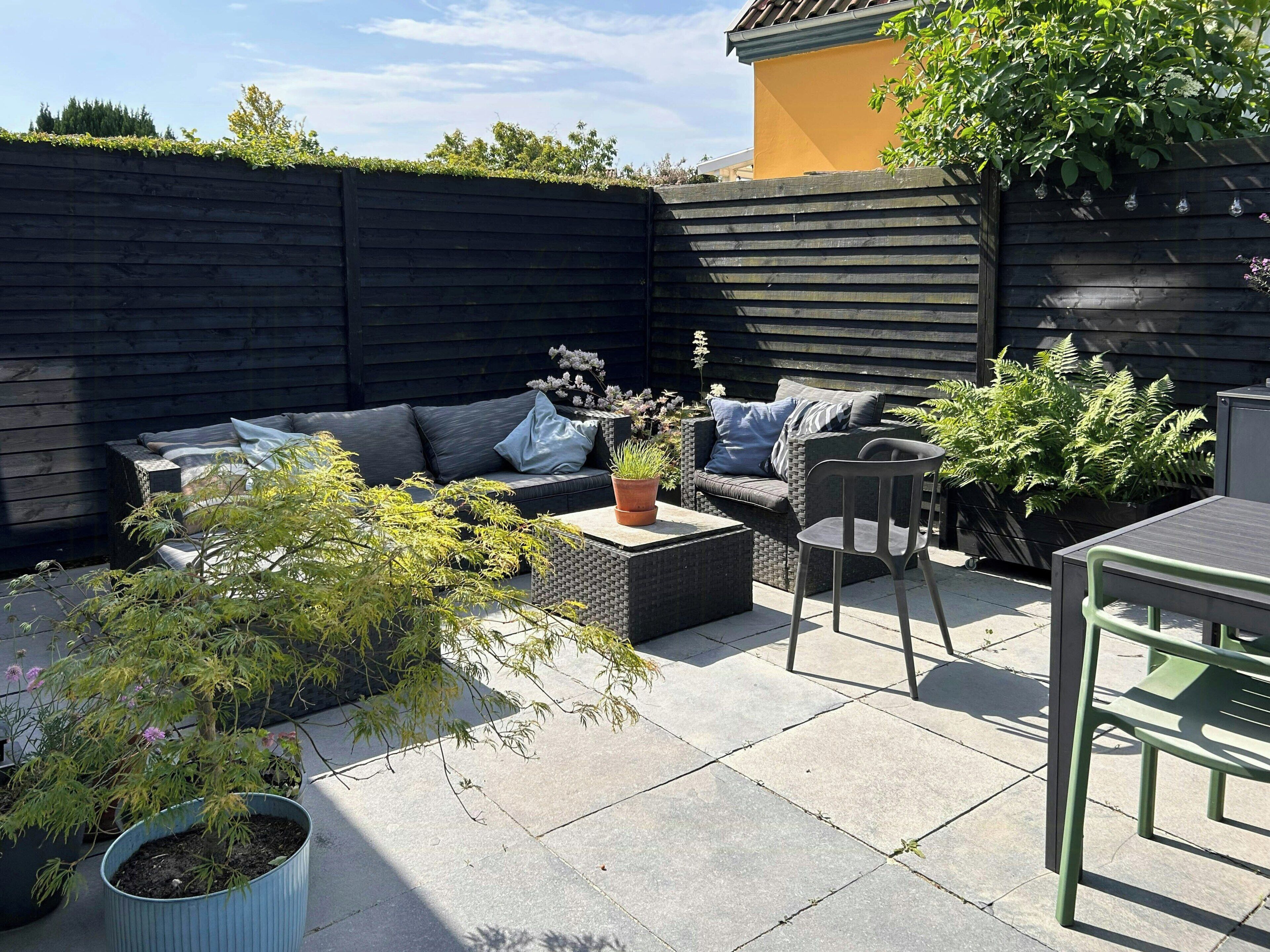 Un patio con pared oscura, mobiliario exterior y plantas en macetas.
