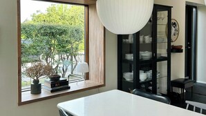 Ett modernt matområde med ett stort fönster, ett vitt bord, svarta stolar, en hängande lampa och ett svart skåp med glasdörrar.