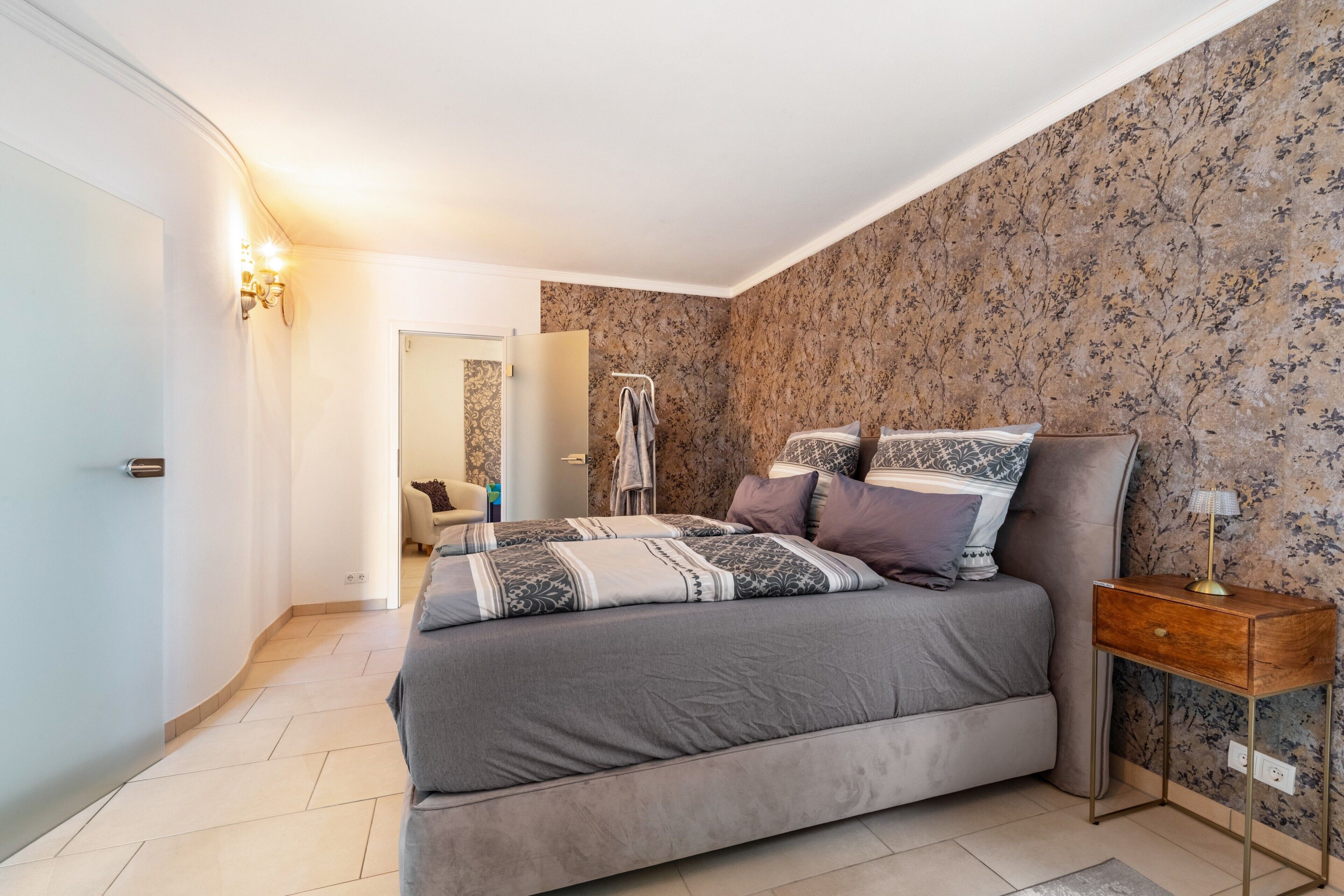 A bedroom with a bed, a nightstand, a lamp, and a bathroom visible in the background.