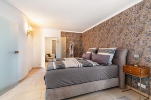 A bedroom with a bed, a nightstand, a lamp, and a bathroom visible in the background.