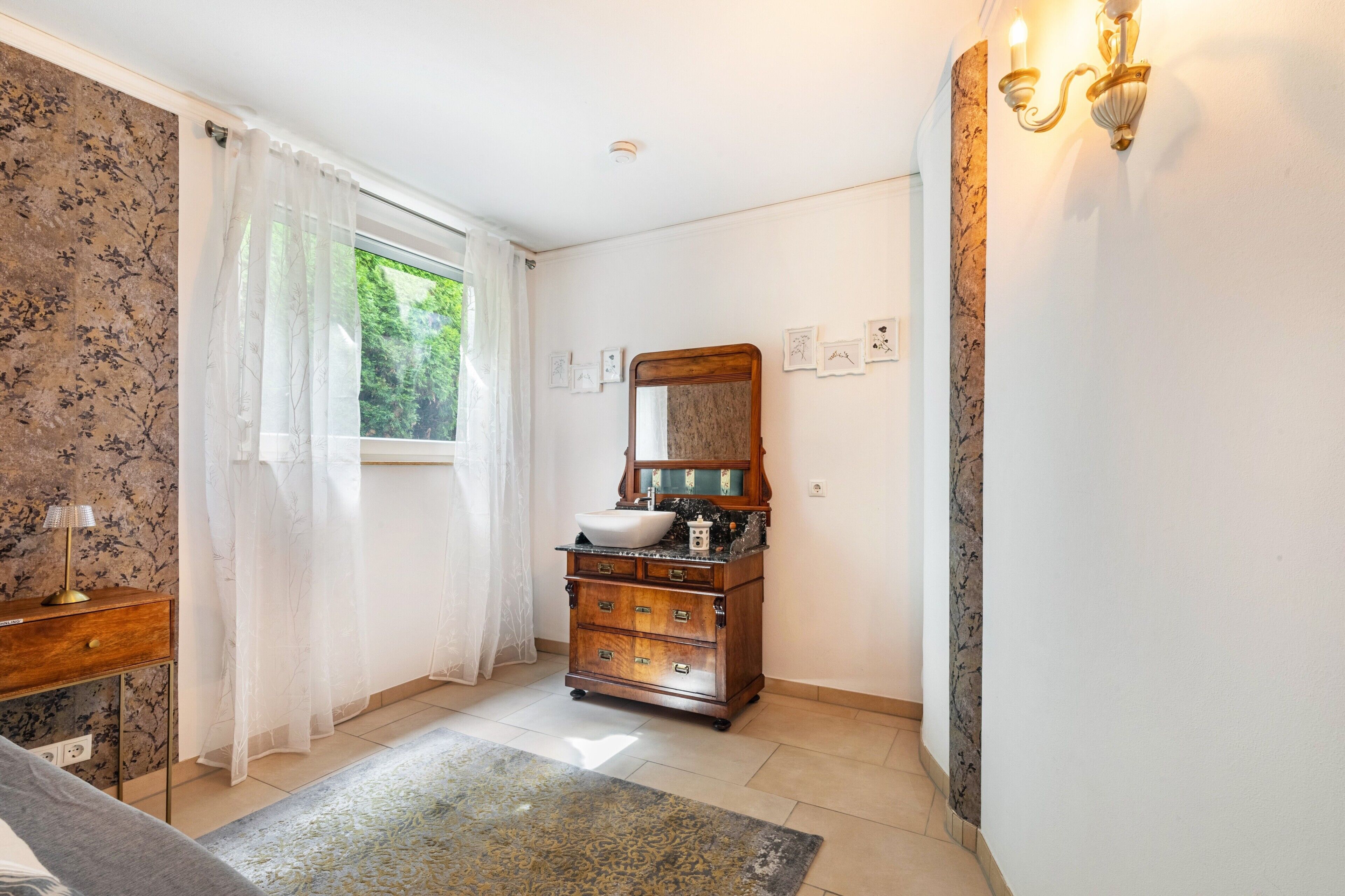A bathroom with a wooden vanity, a large mirror, and a window with sheer curtains.