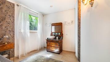 A bathroom with a wooden vanity, a large mirror, and a window with sheer curtains.