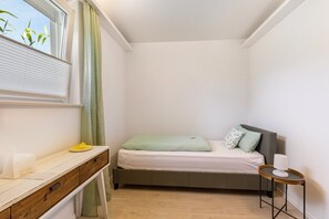 A small, neatly arranged bedroom with a single bed, a bedside table, a small side table, and a window with curtains.