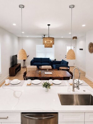 Eine moderne Küche mit Kochinsel, einem Essbereich mit Holztisch sowie ein Wohnbereich mit blauem Sofa und zwei Pendelleuchten aus Rattan.