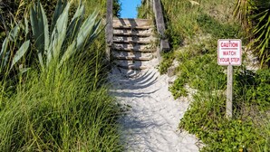 Uma trilha de areia que leva a uma escada de madeira, com uma placa de aviso.