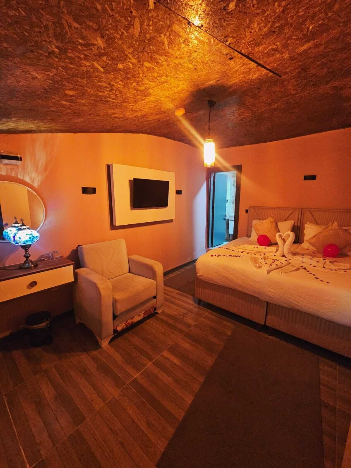Une chambre avec un plafond en bois, un lit avec des oreillers et une couverture, une chaise, un bureau avec une lampe et un téléviseur fixé au mur.