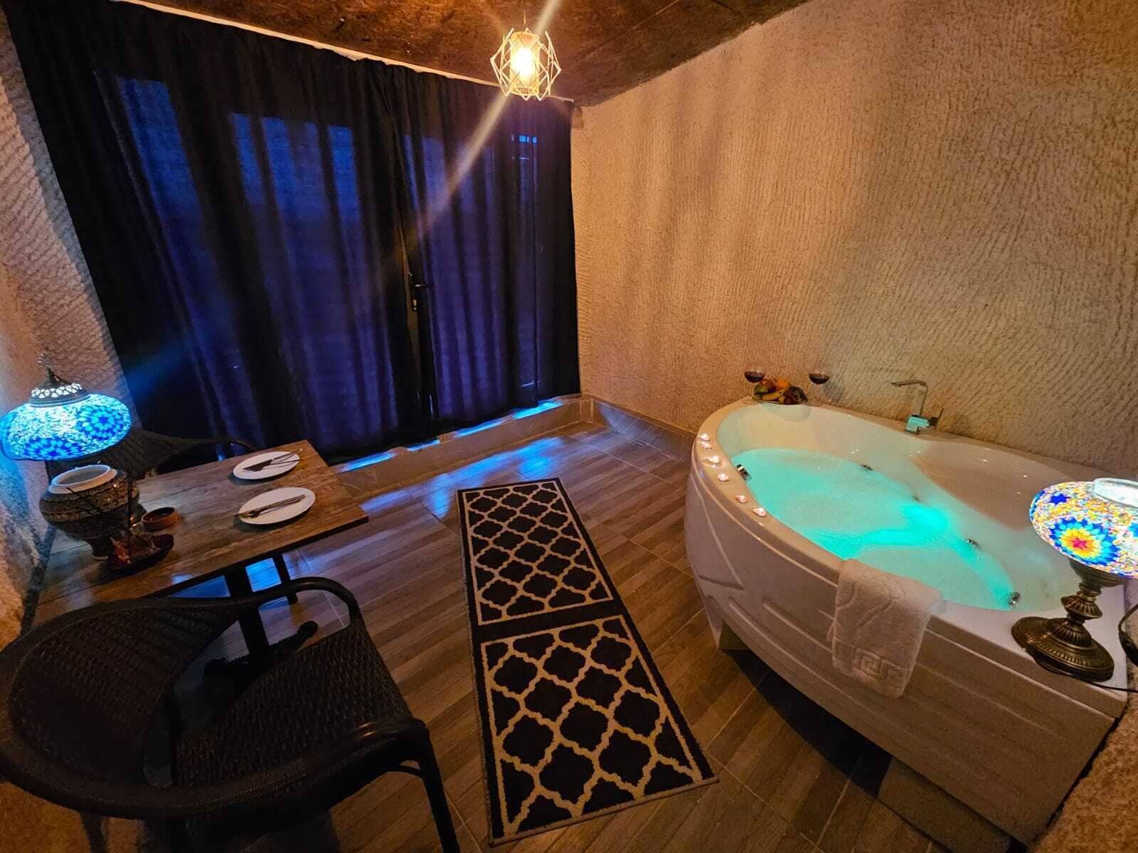 A room with a wooden floor, a bathtub with lights, a table with plates, a chair, and a lamp.
