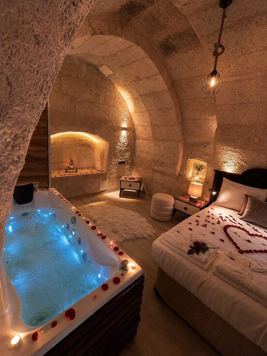 Ein gemütliches Schlafzimmer mit Steinsäulen, einer großen, beleuchteten Badewanne, einem ordentlich bezogenen Bett und einem kleinen Nachttisch mit Lampe.
