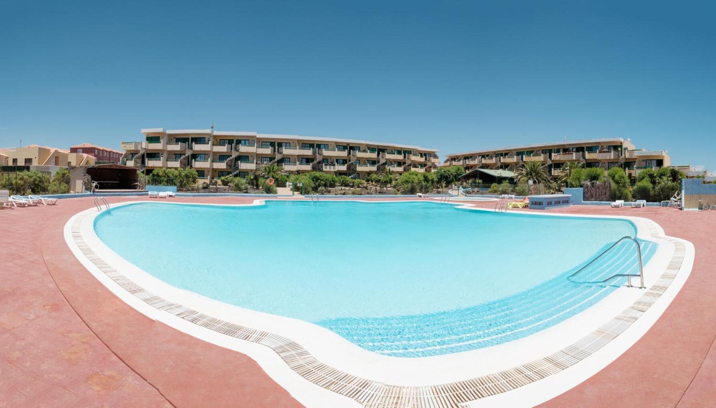 A swimming pool with a curved edge, surrounded by a red deck. In the background, there are multi-story buildings with balconies.