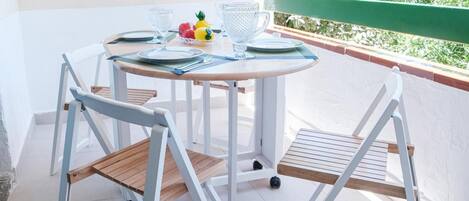 Une table ronde entourée de chaises, un petit saladier rempli de fruits et de la verrerie disposés sur un balcon doté d’une rambarde verte.