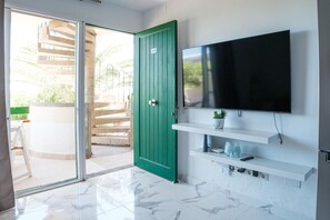 Habitación con puerta corrediza de vidrio que da a un espacio exterior, una puerta verde, televisor montado y estantes blancos con una planta y un control remoto.