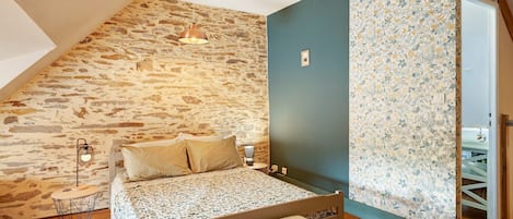 A bedroom with a stone wall, a bed with a floral bedspread, a nightstand with a lamp, and a wooden floor.