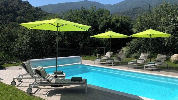 Una piscina amb gandules i ombrelles verdoses, envoltada de vegetació i turons.