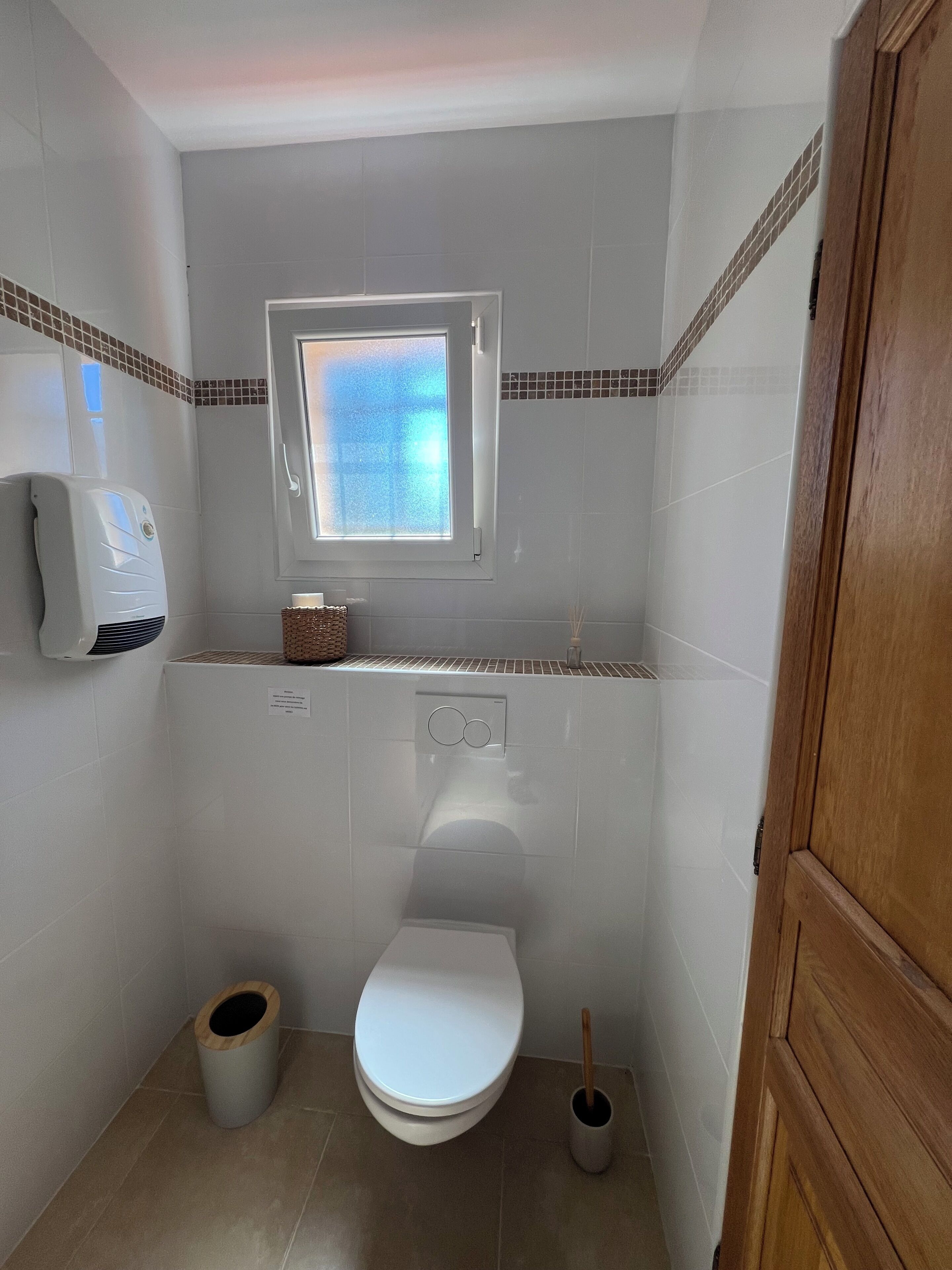 Een kleine badkamer met toilet, raam, handdroger en afvalbak.