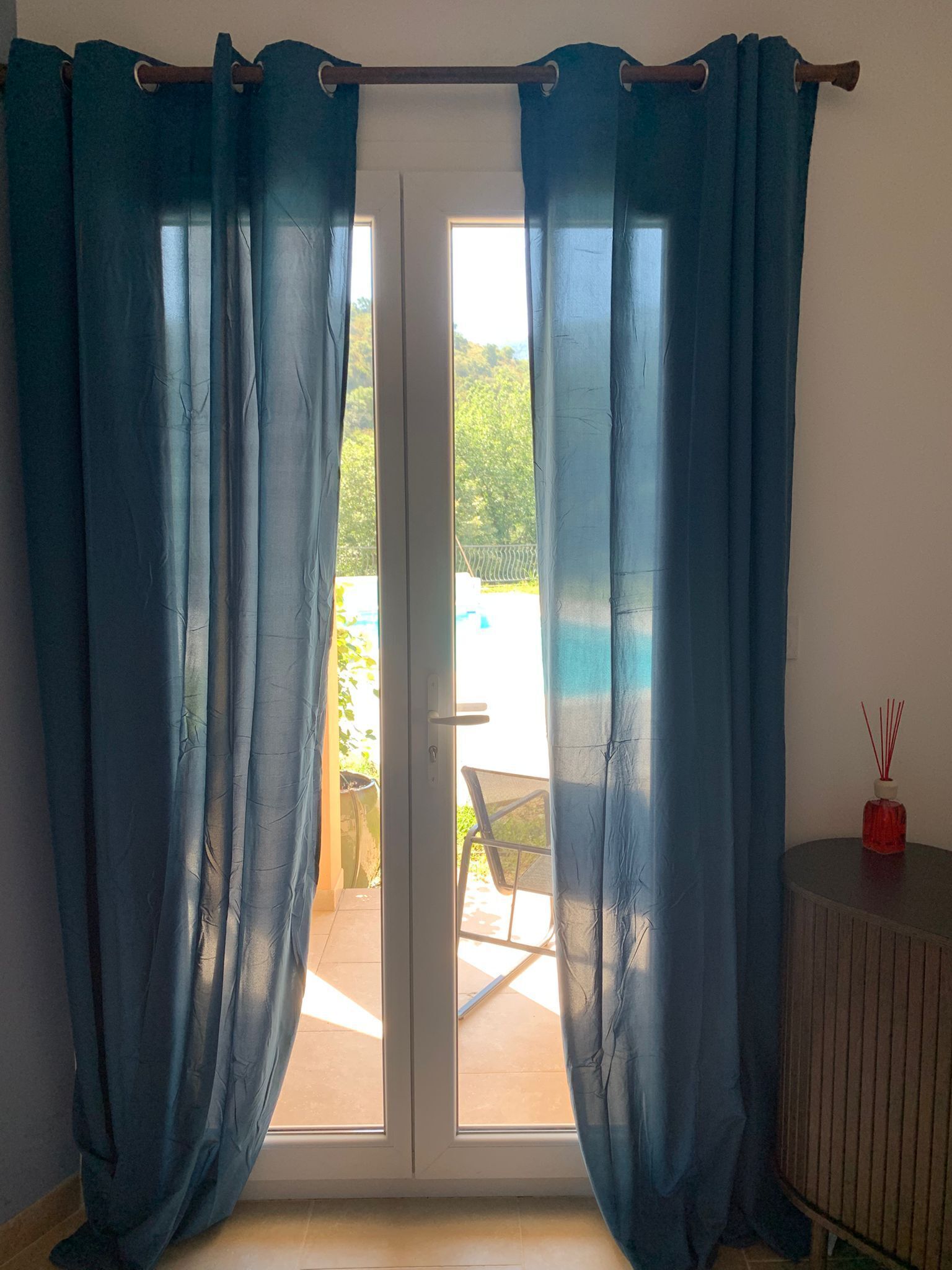 Une pièce dotée d’une porte-fenêtre coulissante, d’une petite table sur laquelle se trouve une bouteille rouge, et qui offre une vue sur un espace extérieur aménagé avec une chaise et de la verdure.