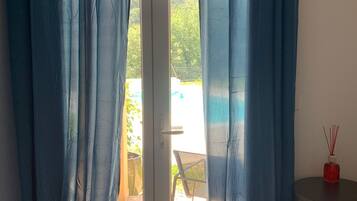 Une pièce dotée d’une porte-fenêtre coulissante, d’une petite table sur laquelle se trouve une bouteille rouge, et qui offre une vue sur un espace extérieur aménagé avec une chaise et de la verdure.