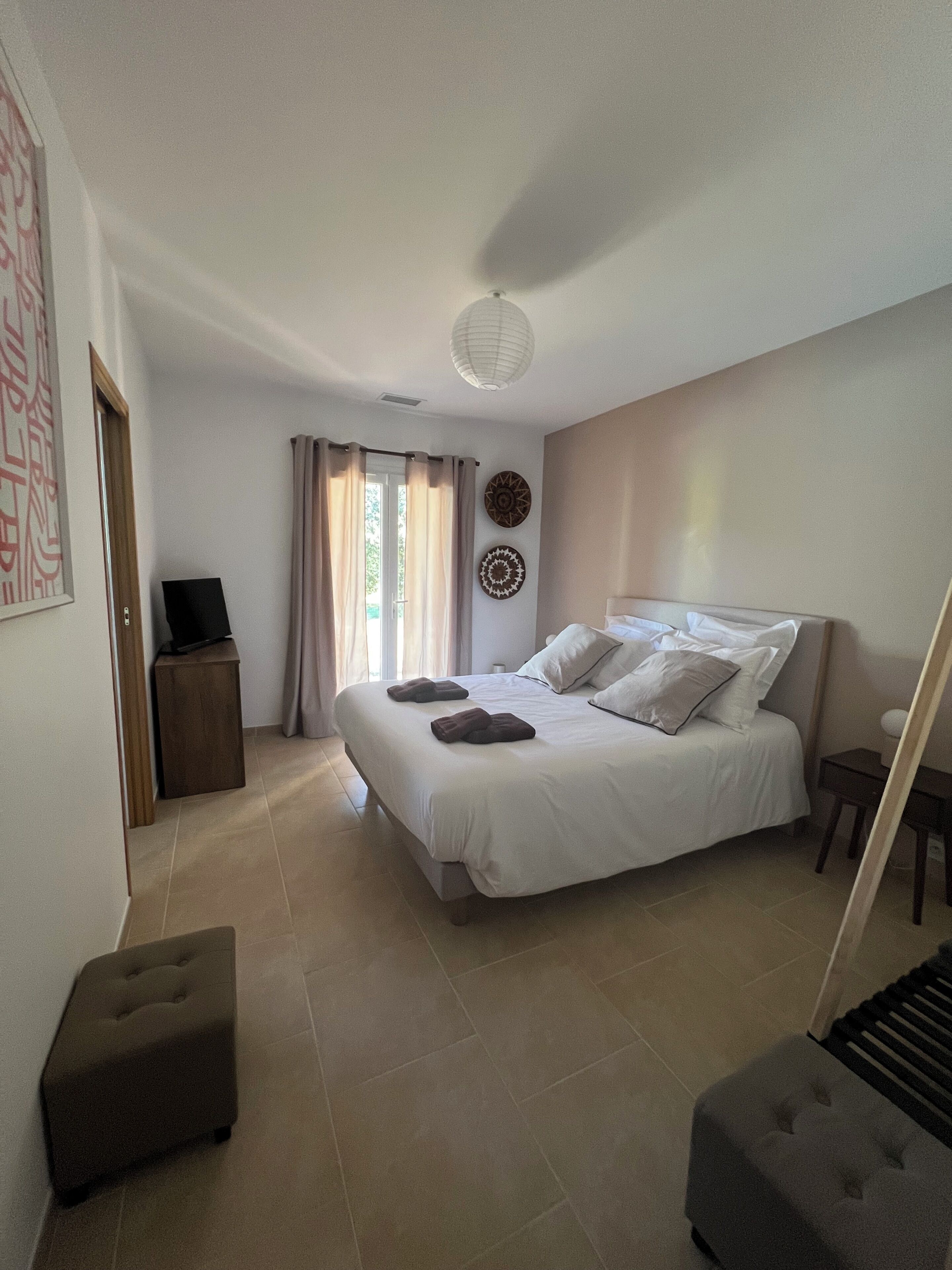 Une chambre à coucher avec un lit, une table de chevet, une chaise, un téléviseur et une fenêtre avec des rideaux.