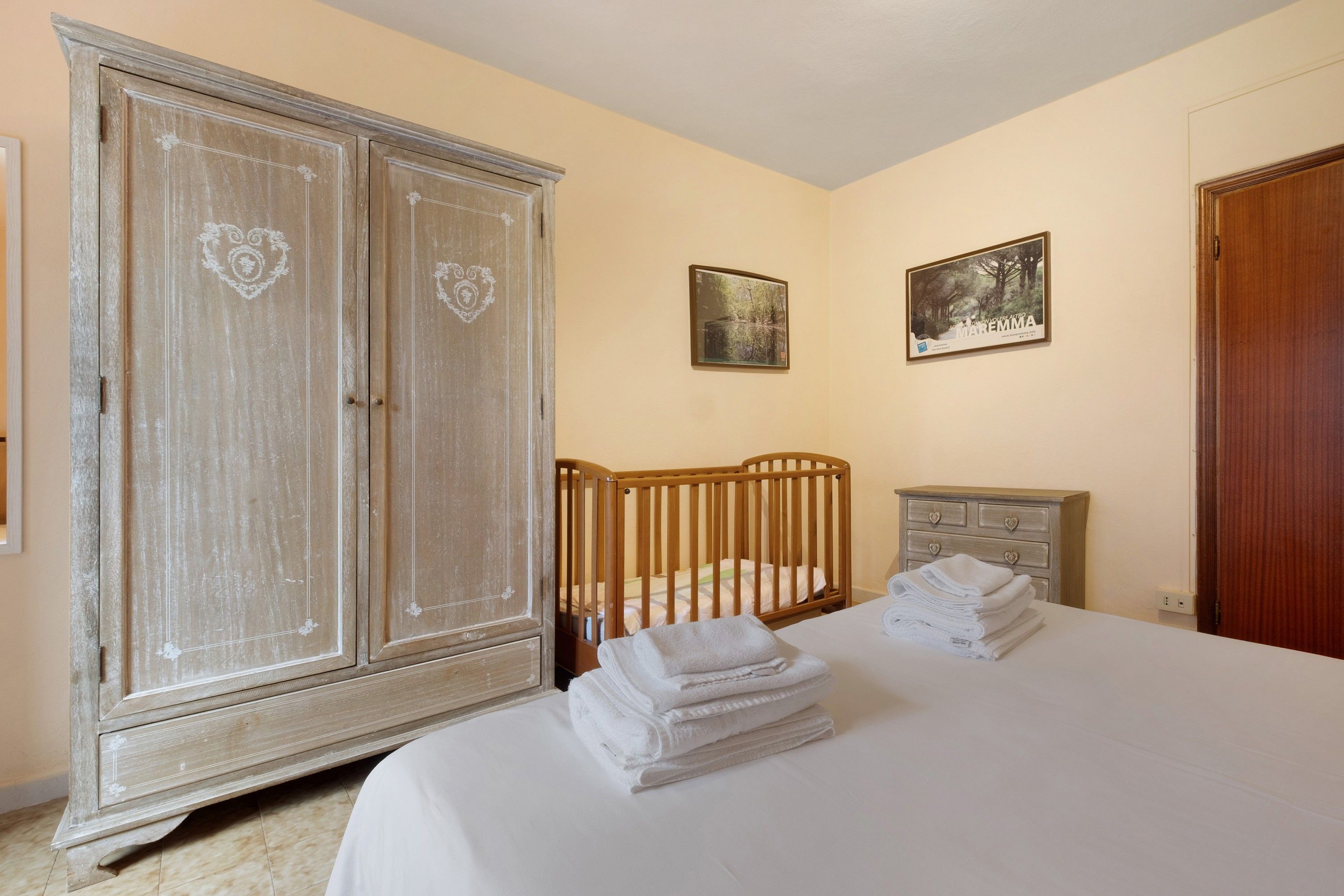 Un dormitorio con un armario grande, una cuna, una mesita de noche con cajón y una cama con toallas dobladas cuidadosamente.
