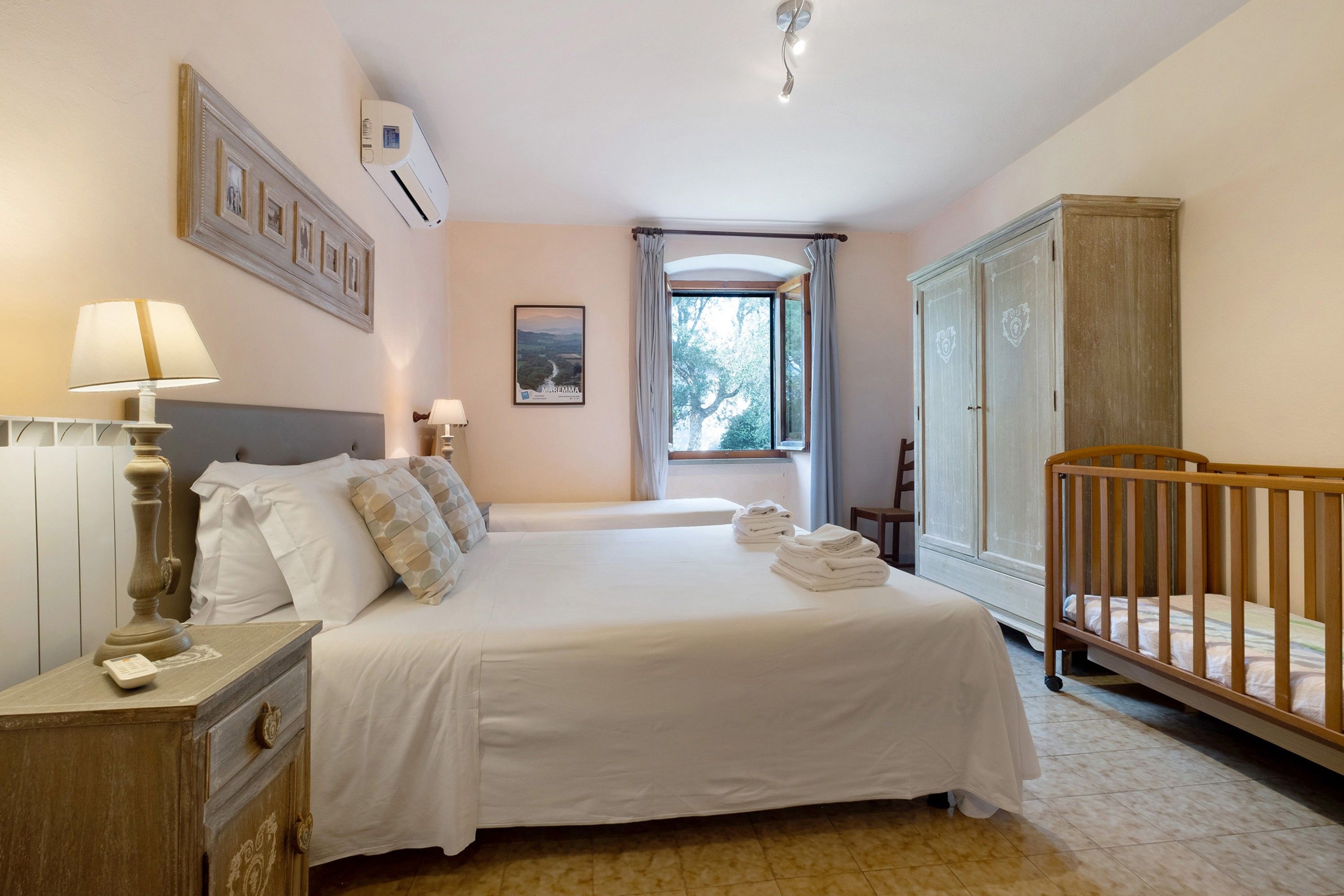 A bedroom with a bed, a nightstand, a dresser, a crib, a window with curtains, and a framed picture on the wall.